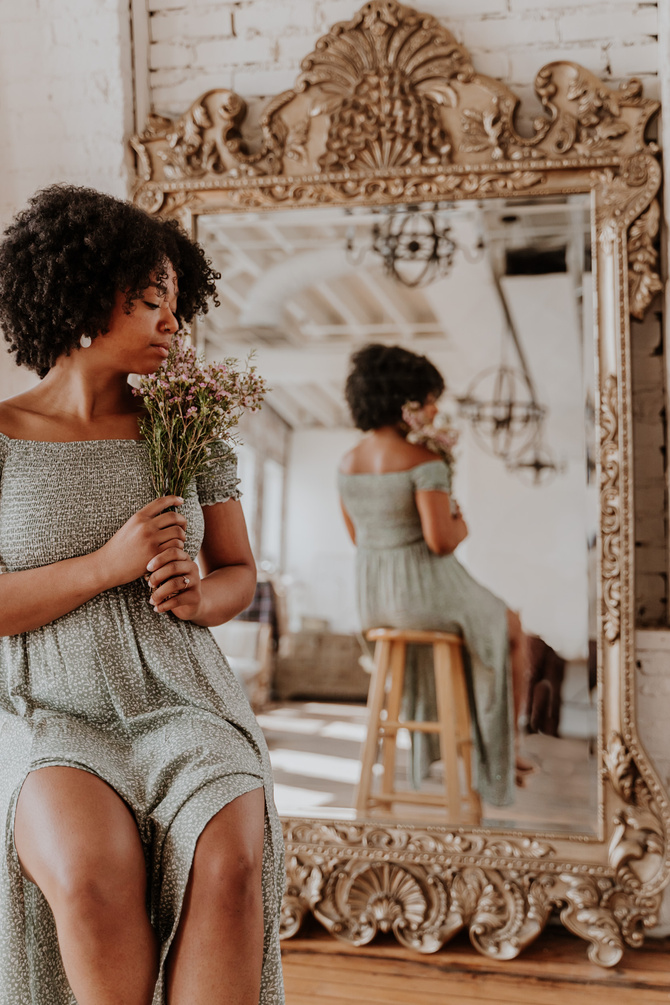 A woman in a green dress holding wildflowers beside an antique mirror