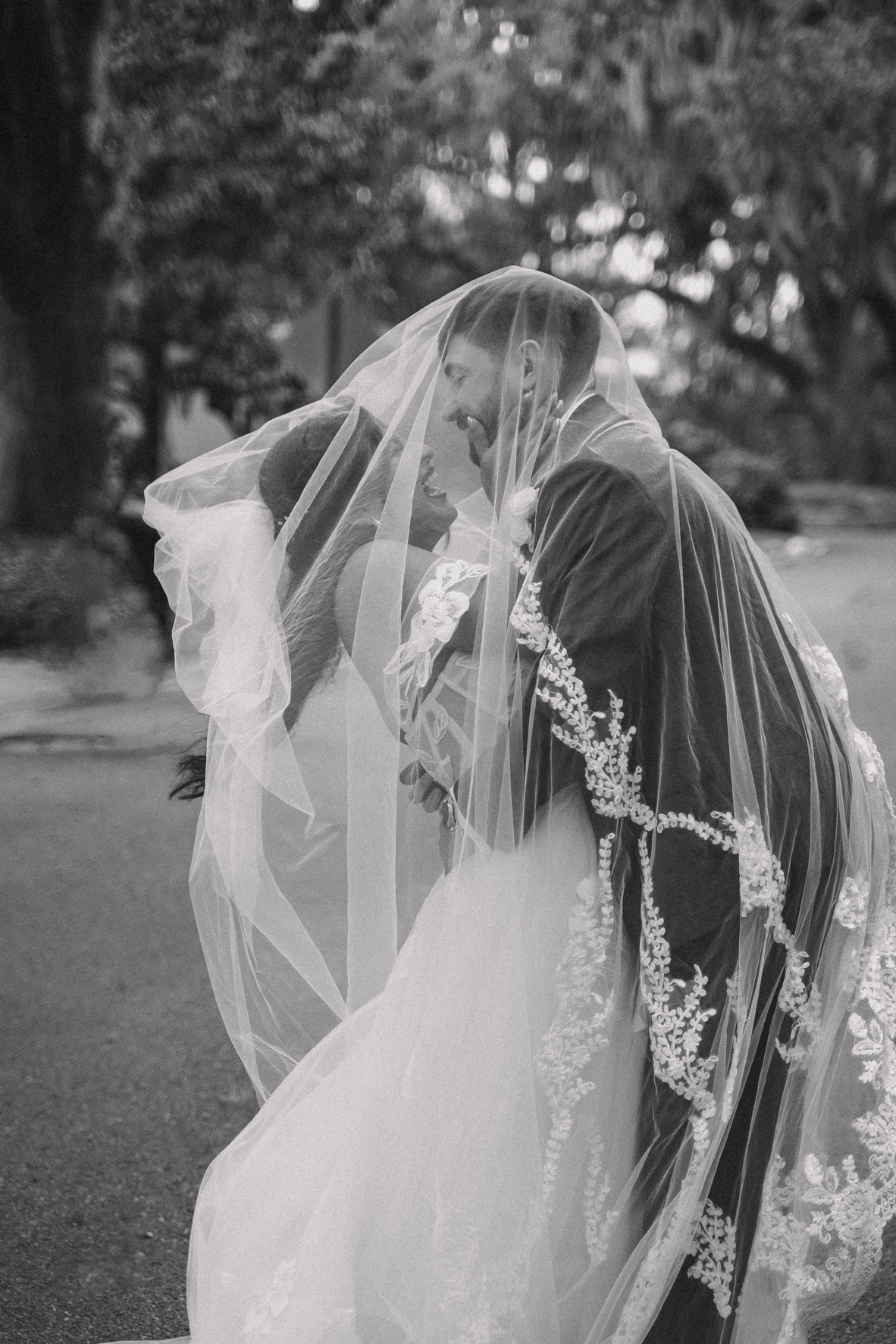 A bride and groom embraced beneath a long lace veil, black and white